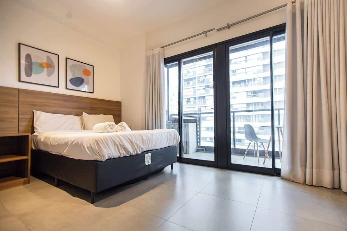 Una habitación de hotel moderna con una cama grande, un cabecero de madera, un balcón con una silla y dos cuadros abstractos en la pared.