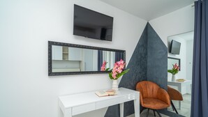 Ein modernes Zimmer mit weißem Schreibtisch, einem großen Spiegel, einem an der Wand montierten Fernseher und einem Sessel.
