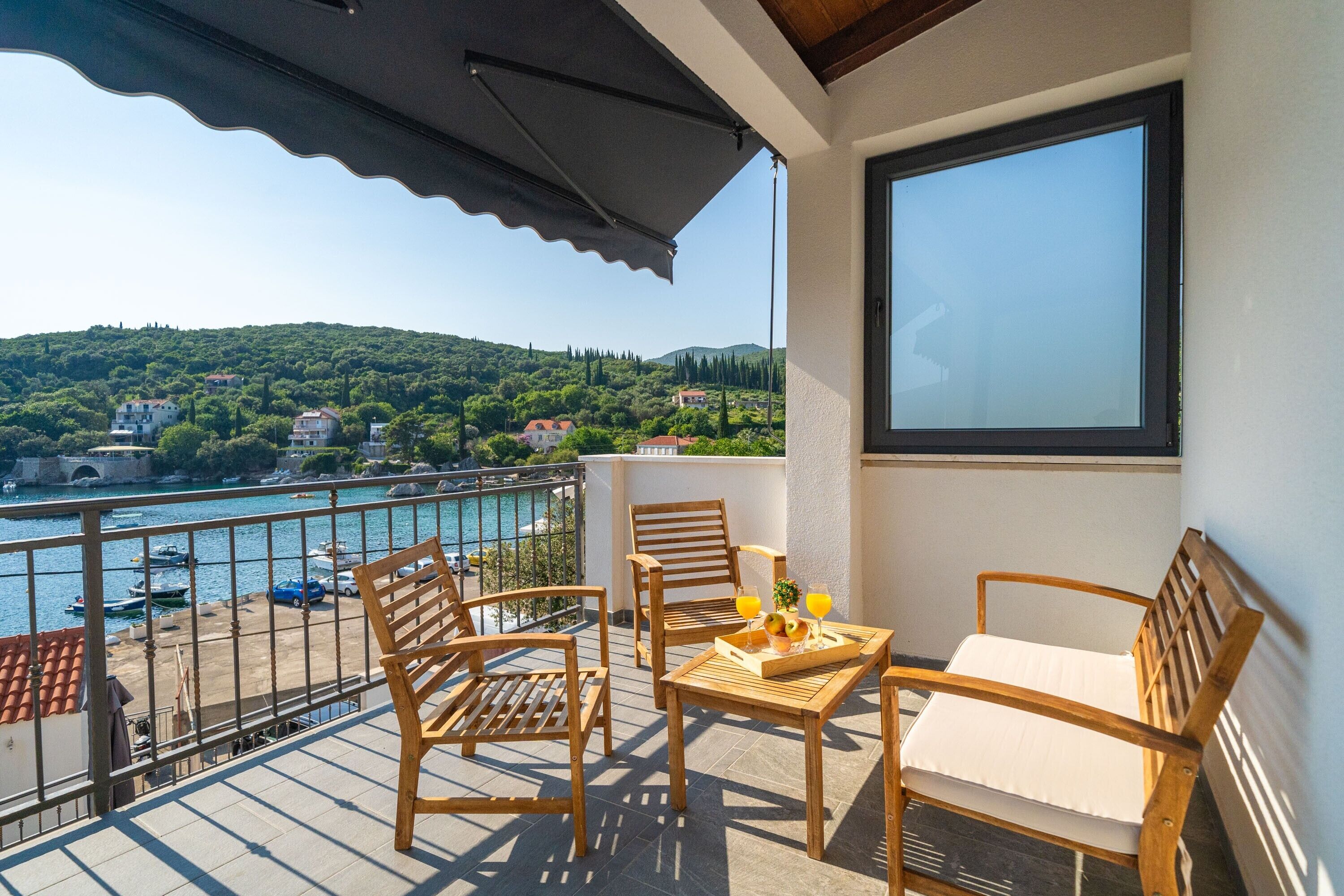 A balcony with wooden furniture, a table with a tray of fruit and drinks, and a view of a coastal town and hills.