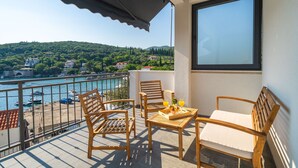 Ein Balkon mit Holzmöbeln, einem Tisch mit Obst und Getränken sowie Blick auf eine Küstenstadt und Hügel.