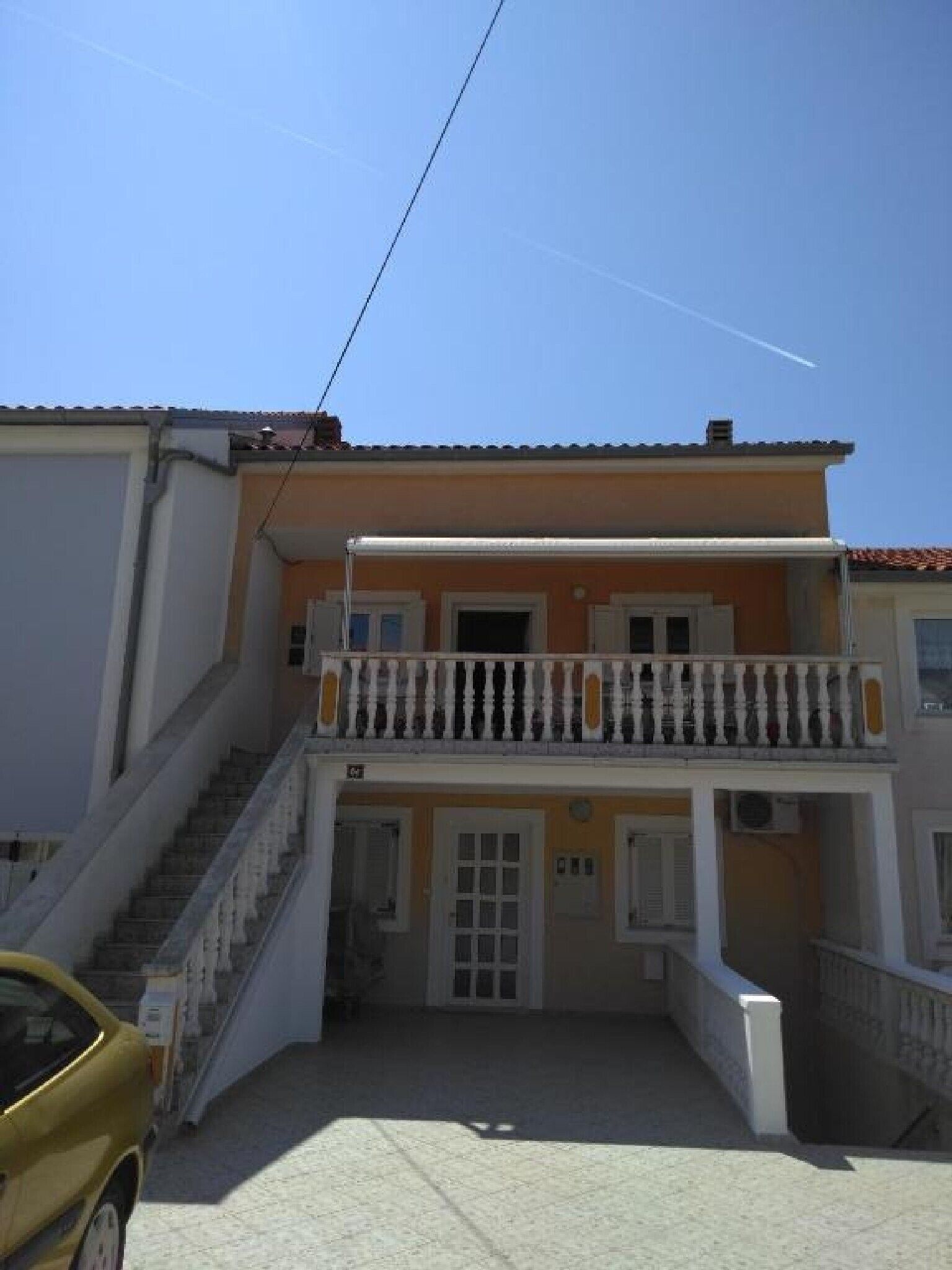 A two-story building with a balcony, a staircase leading to the upper floor, and a car parked in the driveway.