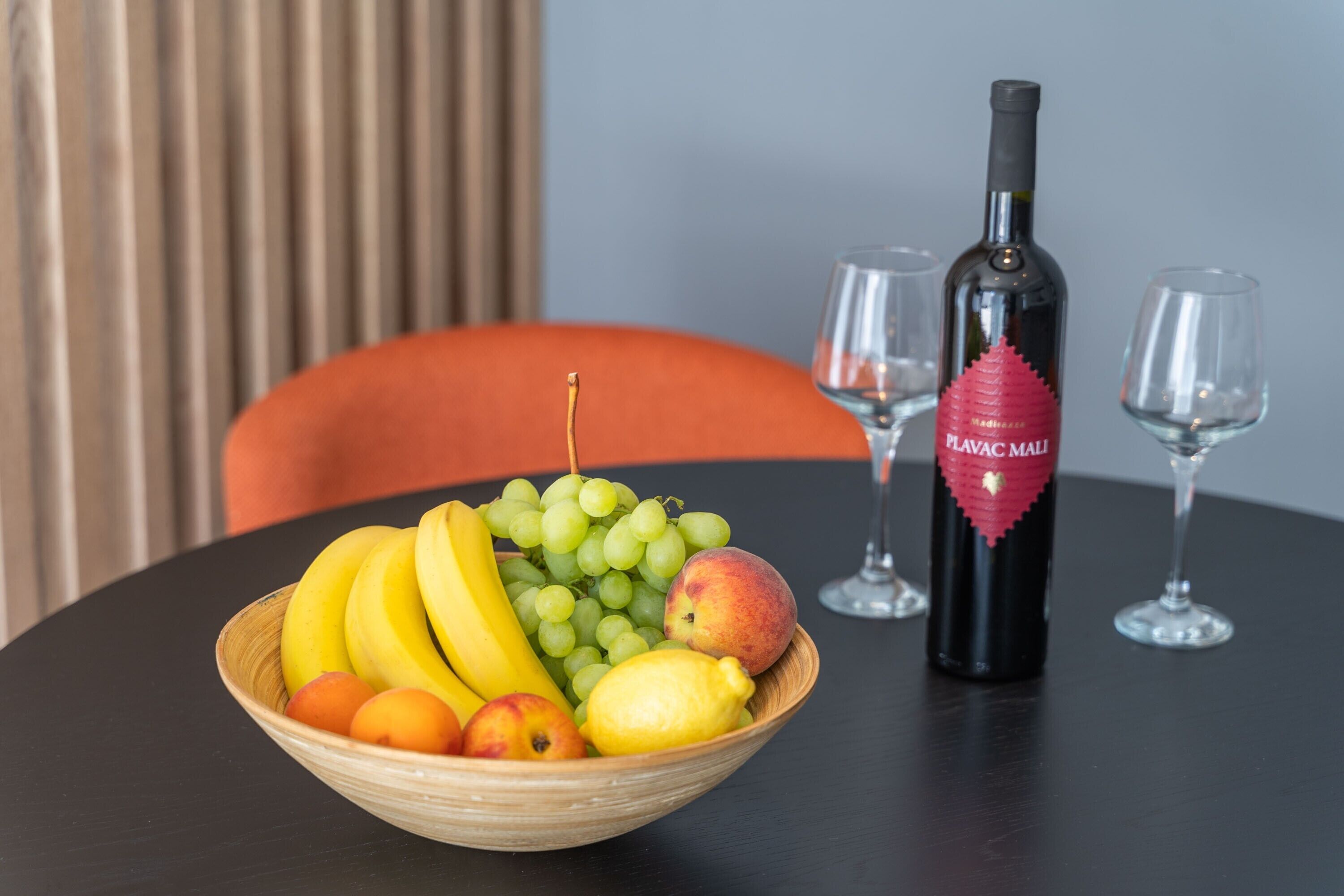 Un tavolo con una ciotola di frutta contenente banane, uva, pesche e un limone. C'è anche una bottiglia di vino e due bicchieri sul tavolo.