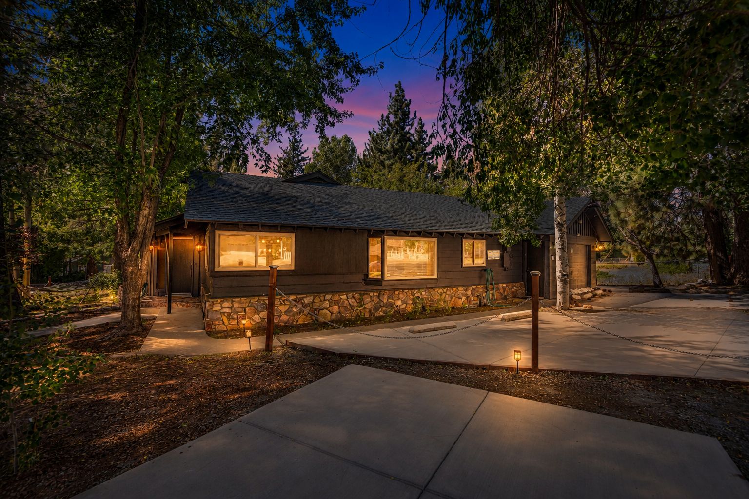 A rustic cabin with a stone foundation, large windows, and a well-lit exterior at dusk.
