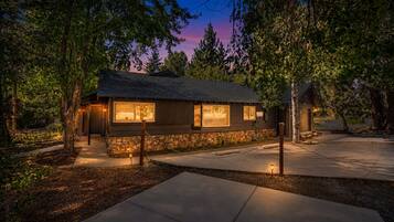 A rustic cabin with a stone foundation, large windows, and a well-lit exterior at dusk.