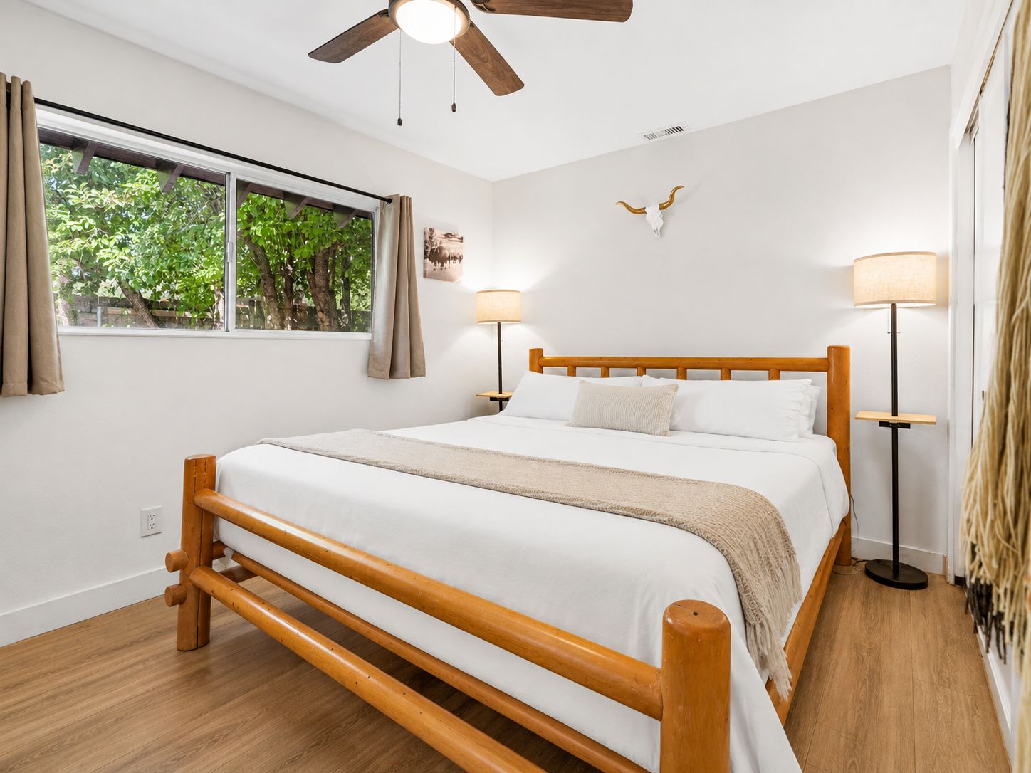 Un dormitorio con una cama de madera, dos lámparas de noche, un ventilador de techo, una ventana con cortinas y un adorno en la pared.
