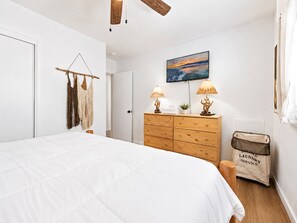 Quarto com cama, cômoda de madeira, duas luminárias na parede, um quadro e um cesto de roupa.