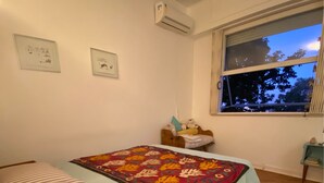 A bedroom with a bed, a bedside table, an air conditioning unit, a window with a view of trees, and a wall-mounted clock. - Charming and peaceful Santa Teresa (Santa Teresa Rio de Janeiro)