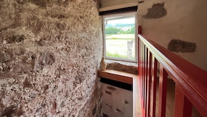 A rustic room with exposed brick walls, a wooden staircase, and a window offering a view of a green landscape.