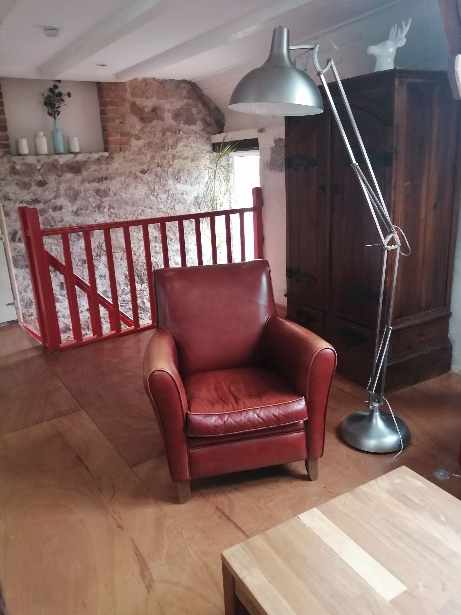 A red leather armchair, a wooden table, and a floor lamp in a room with a rustic brick wall and a red metal gate.