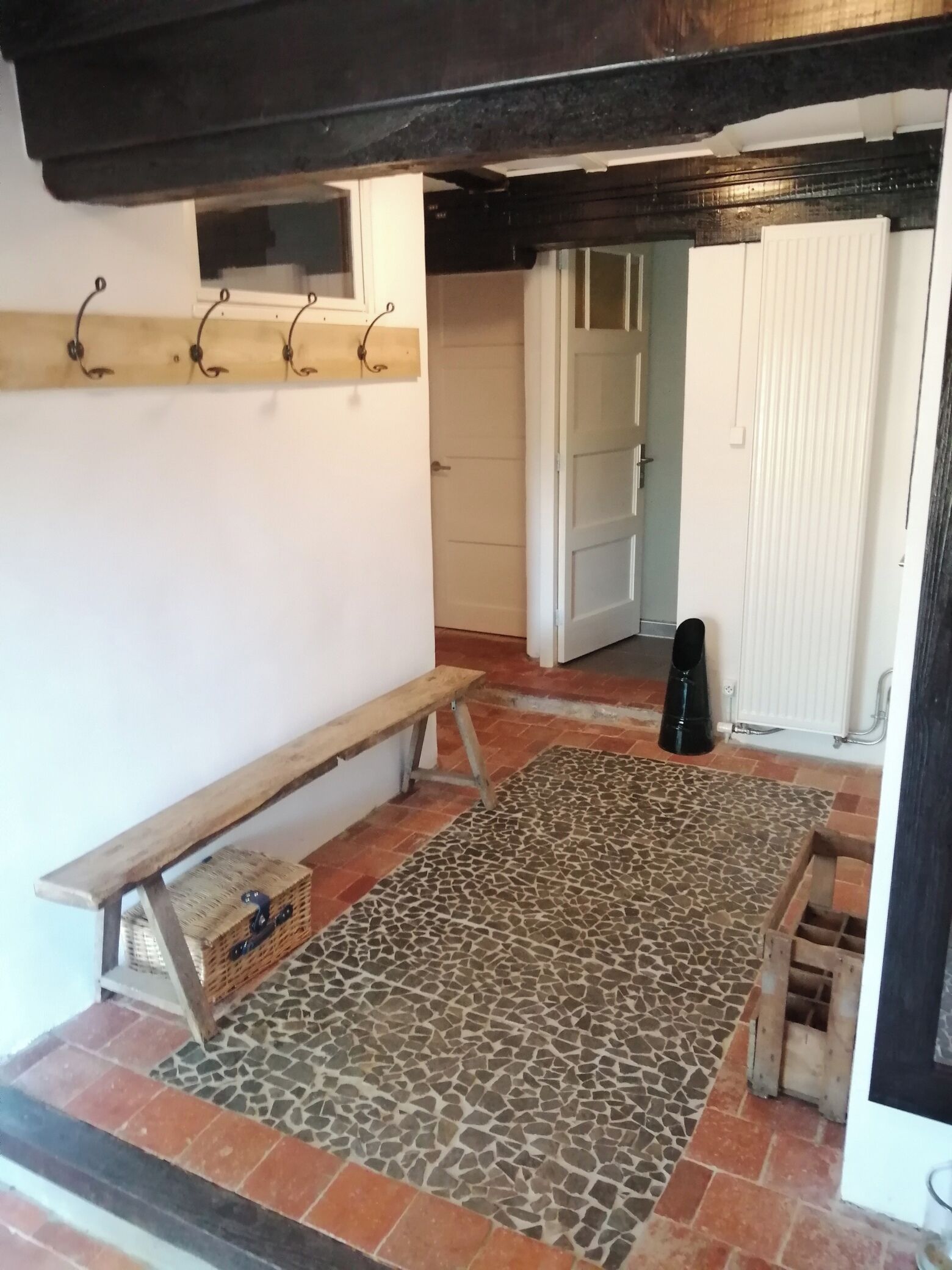 Een kamer met een bank, een kamerbreed tapijt, een houten bank met manden, en een deur die naar een andere kamer leidt.