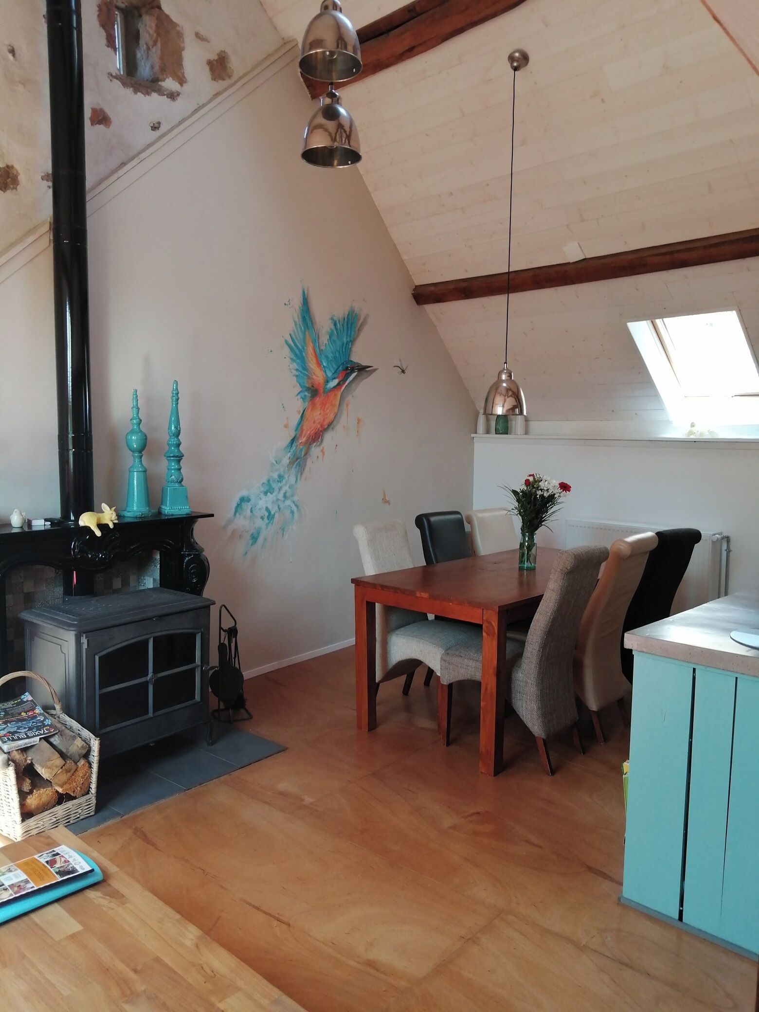 Un salon chaleureux avec un plancher en bois, une cheminée, une table à manger avec des chaises et une fresque murale représentant un oiseau.