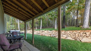Een overdekt terras met een tafel en stoelen, een stenen muur en een gazon met bomen.