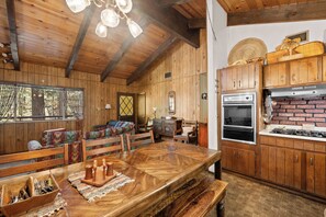 A rustic cabin interior with a wooden dining table, benches, a kitchen with a brick wall, and a large window overlooking a forest. - Gallagher's West (Big Bear Lake)