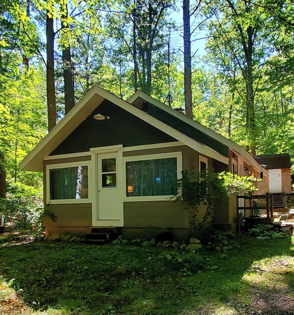 A small cabin with a gabled roof, a front door, and windows, surrounded by trees and greenery.