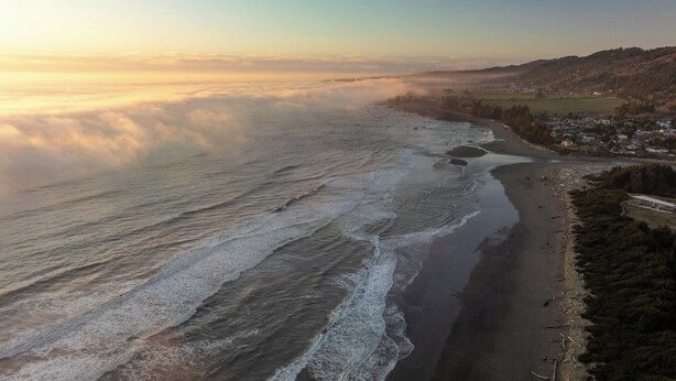 Vista aérea de uma área costeira com praia, ondas e uma cidade ao fundo.