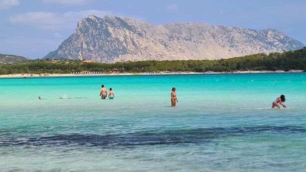 Une plage à l’eau turquoise et limpide, où l’on peut voir des gens nager et se promener, avec une montagne en arrière-plan.