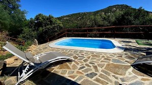 Une piscine en pierre entourée de chaises longues, elle-même bordée d’une terrasse en pierre et d’une clôture en métal, avec une chaîne de montagnes en arrière-plan.