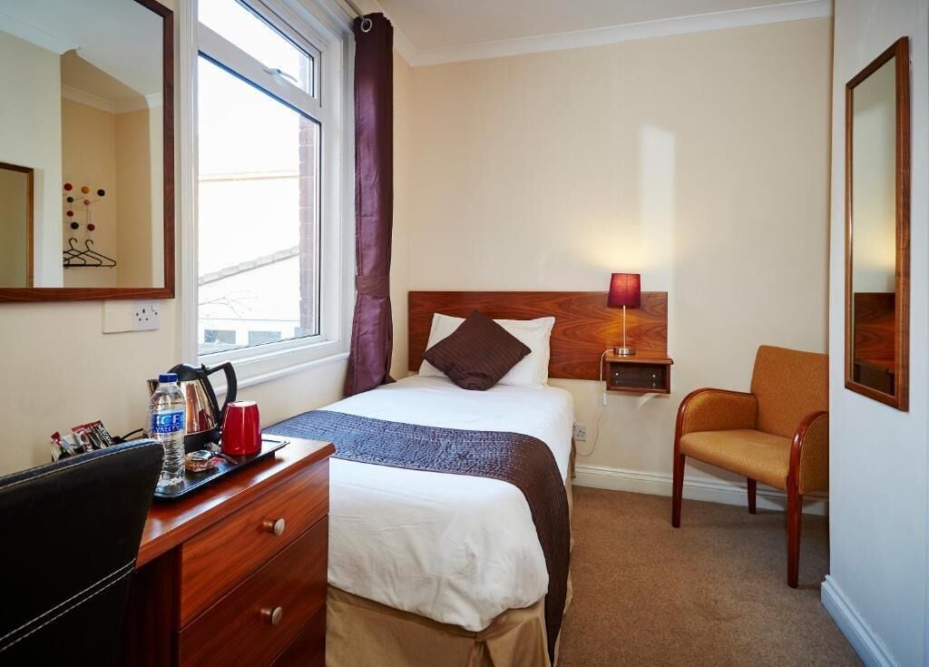A hotel room with a bed, a desk with a chair, a lamp, and a window with curtains.