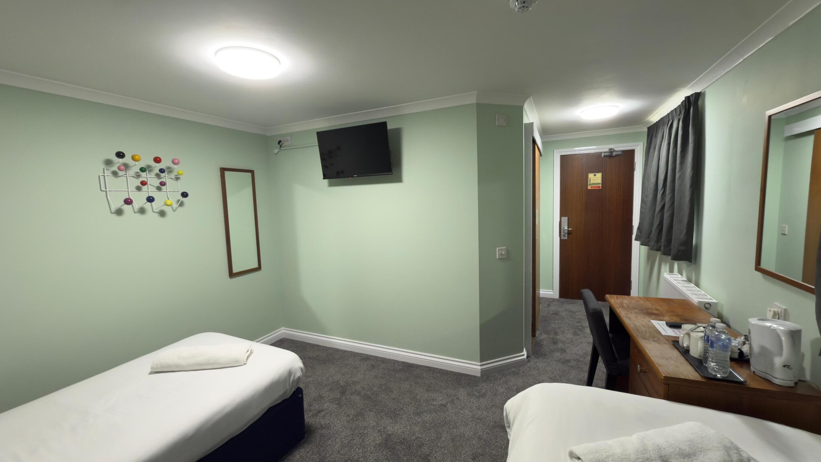 Een hotelkamer met twee eenpersoonsbedden, een bureau met stoel, een flatscreen televisie aan de muur en een klein tafeltje met waterkoker en kopjes.
