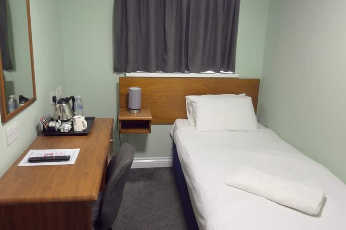Ein Hotelzimmer mit einem Bett, einem Schreibtisch, einem Stuhl, einem Nachttisch, einer Lampe, einem Spiegel und einem Fenster mit Vorhängen.