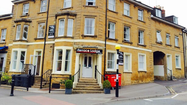 A corner building with a sign for Terrace Lodge Hotel. It has multiple windows, a front entrance with steps, and a small garden area with potted plants. - Terrace Lodge (Yeovil)