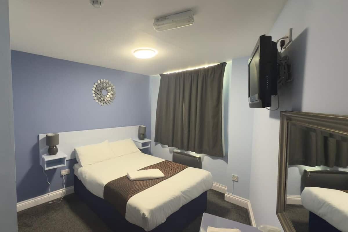 Ein Hotelzimmer mit Bett, Nachttischen, einem Flachbildfernseher, einem Fenster mit Vorhängen und einem an der Wand befestigten Spiegel.