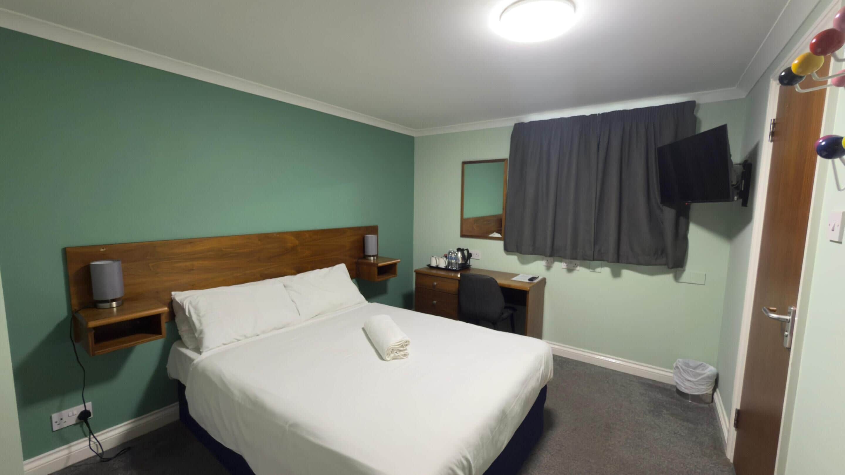 A hotel room with a bed, bedside table, lamp, and a TV mounted on the wall.