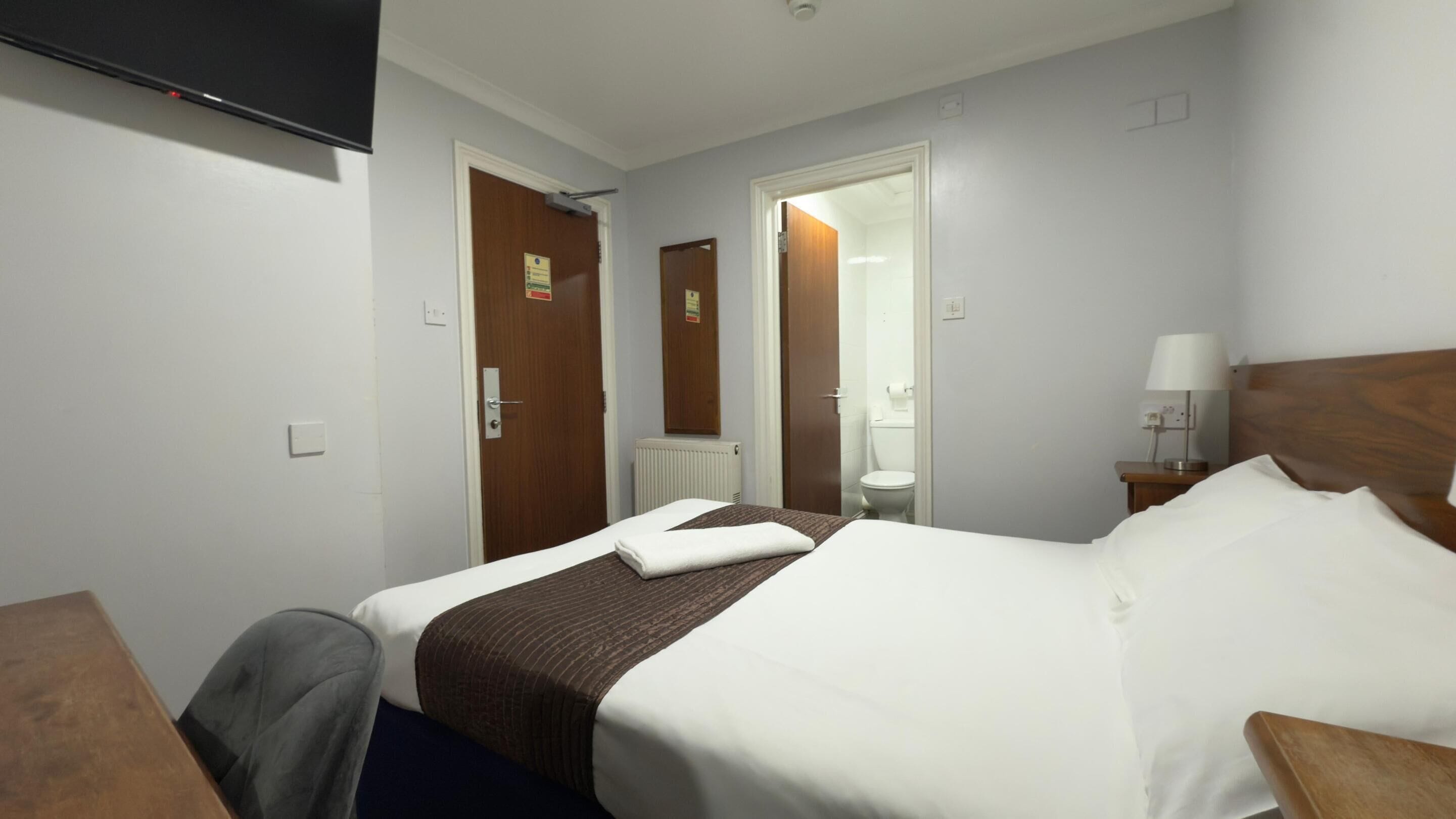 A hotel room with a bed, a desk, a chair, a bedside table with a lamp, and a television mounted on the wall.