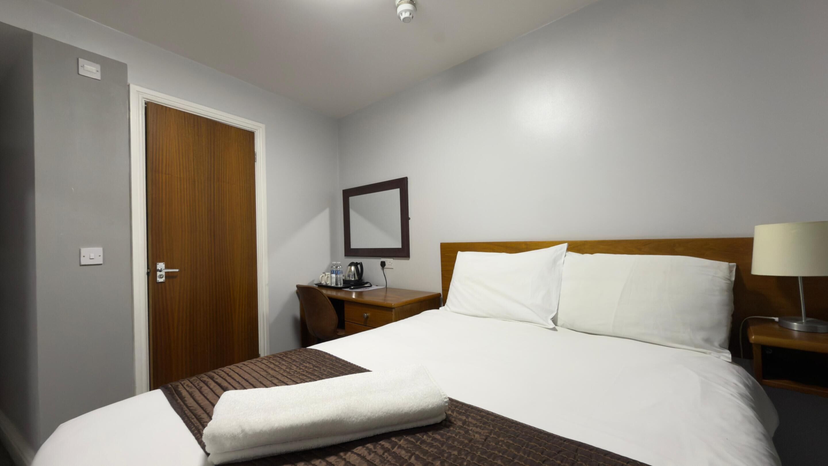 Una habitación de hotel con una cama, una puerta de madera, una mesita de noche con lámpara y un espejo en la pared.