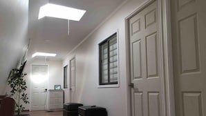 A hallway with white walls, a wooden floor, and a ceiling with recessed lighting. There are two black storage bins and a potted plant.