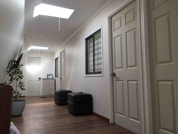 A hallway with white walls, a wooden floor, and a ceiling with recessed lighting. There are two black storage bins and a potted plant.