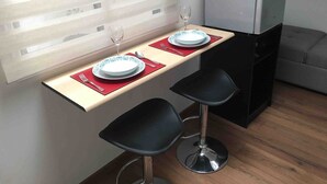 Eine kompakte Küchenzeile mit Kühlschrank, einem kleinen Esstisch mit zwei schwarzen Barhocker und einem Fenster mit Jalousien.