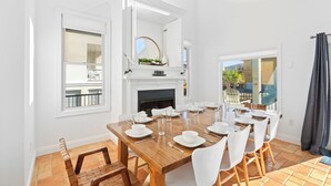 Un coin repas avec une table en bois dressée pour six personnes, des chaises blanches, une cheminée et une vue sur l’extérieur.