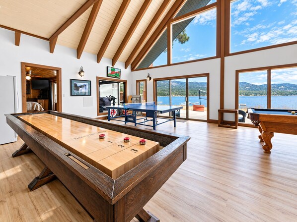 A spacious room with a pool table, a ping pong table, and a billiards table. There are large windows offering a view of a lake and mountains. - Hakuna Matata Lakefront (Big Bear Lake)