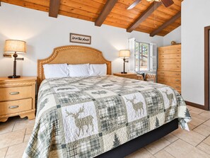 A bedroom with a wooden ceiling, a bed with a quilt, bedside tables, a dresser, and a window with shutters.