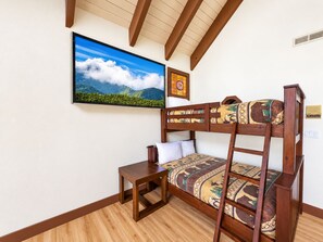 A bunk bed with a patterned mattress, a small wooden table, a framed picture of a mountain landscape, and a window with a view of the outdoors.