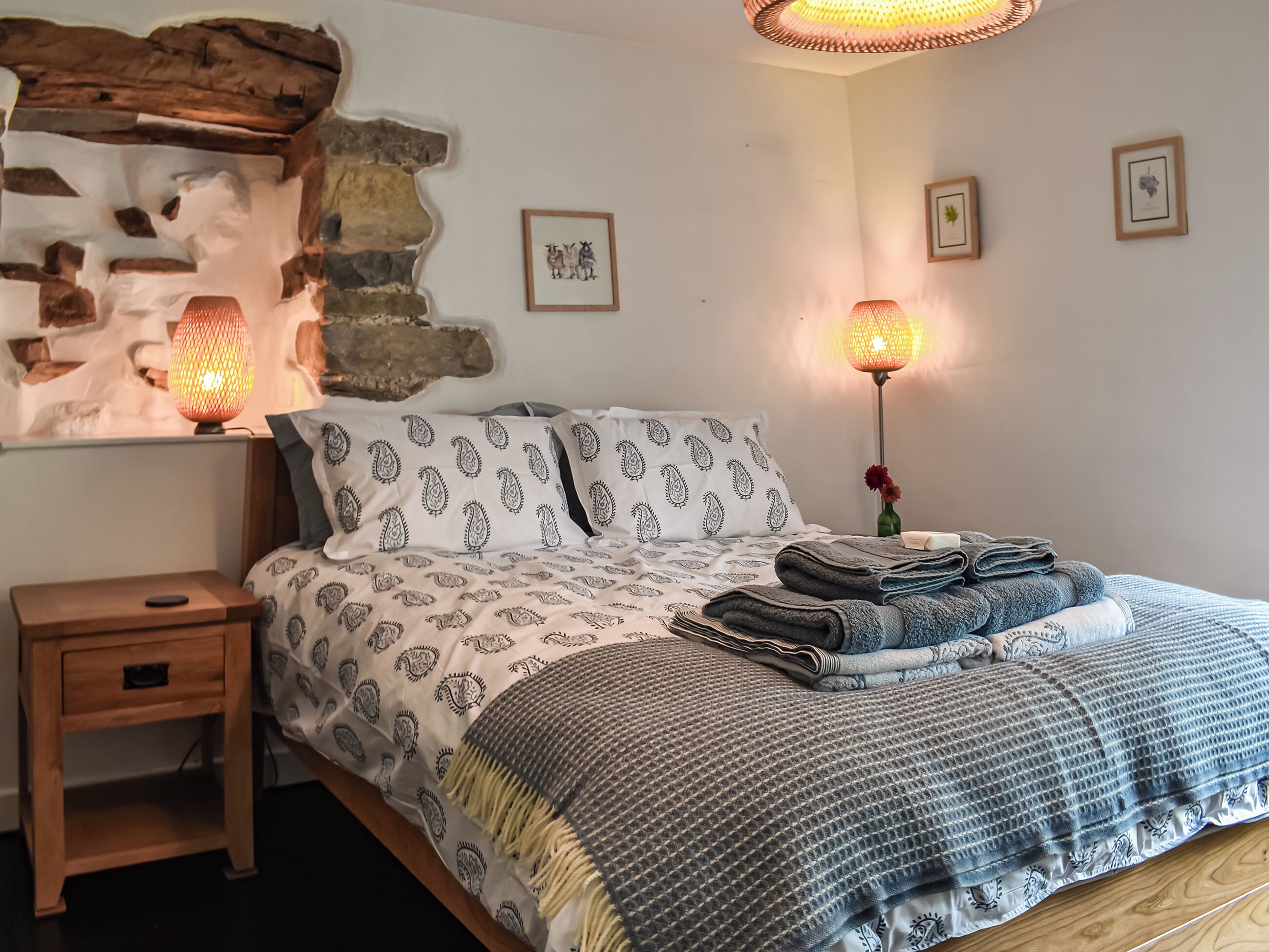 A bedroom with a stone wall, a bed with patterned bedding, two wall-mounted lamps, and a bedside table with a drawer.