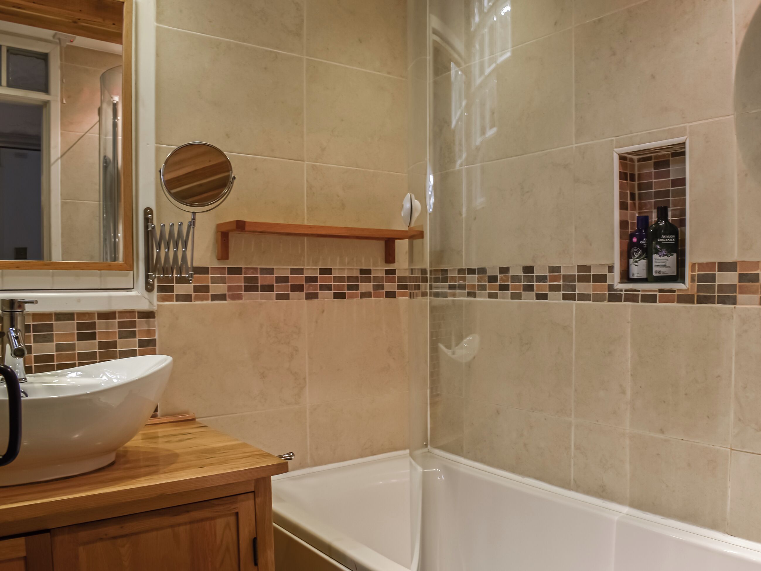 A bathroom with a white bathtub, a wooden vanity with a white sink, and a mirror above the sink. There is a shelf with toiletries and a small window with a view of the outside.