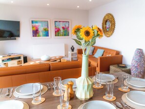 Et spisebord dækket med tallerkener, glas og bestik, en vase med solsikker, og en sofa i baggrunden.
