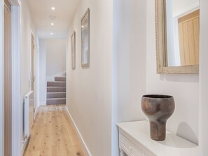 A hallway with wooden flooring, a white wall, a staircase, and a large vase on a white console.