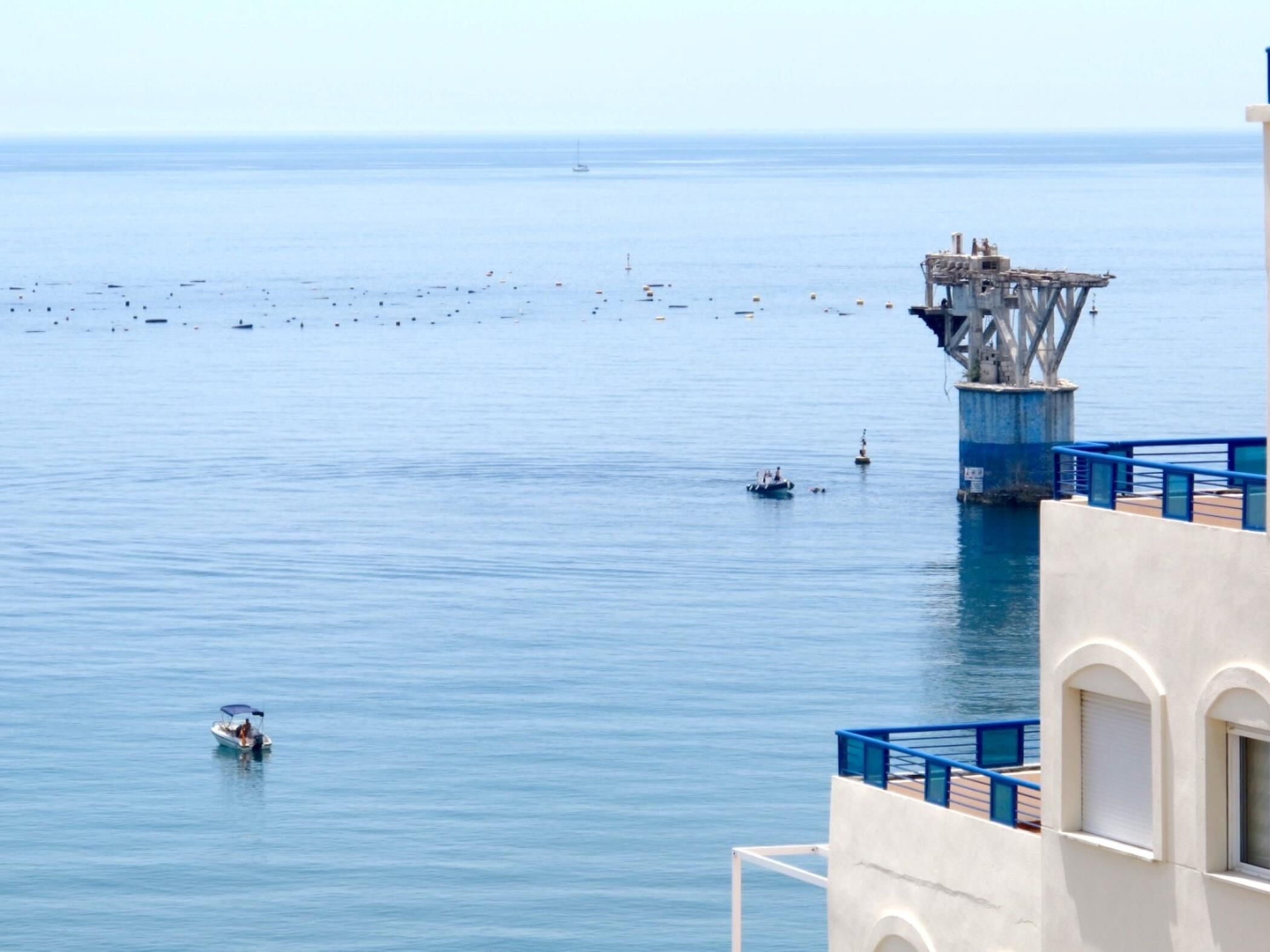 Une vue sur la côte, avec une jetée qui s’avance dans la mer, un bateau au premier plan et un bâtiment doté de balcons donnant sur l’eau.