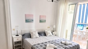 Una cama doble con una colcha estampada, dos mesitas de noche con lámparas, vistas al balcón y dos cuadros colgados en la pared.