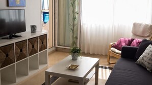 Una sala de estar con televisor, una mesa de centro blanca, un sofá y una planta.