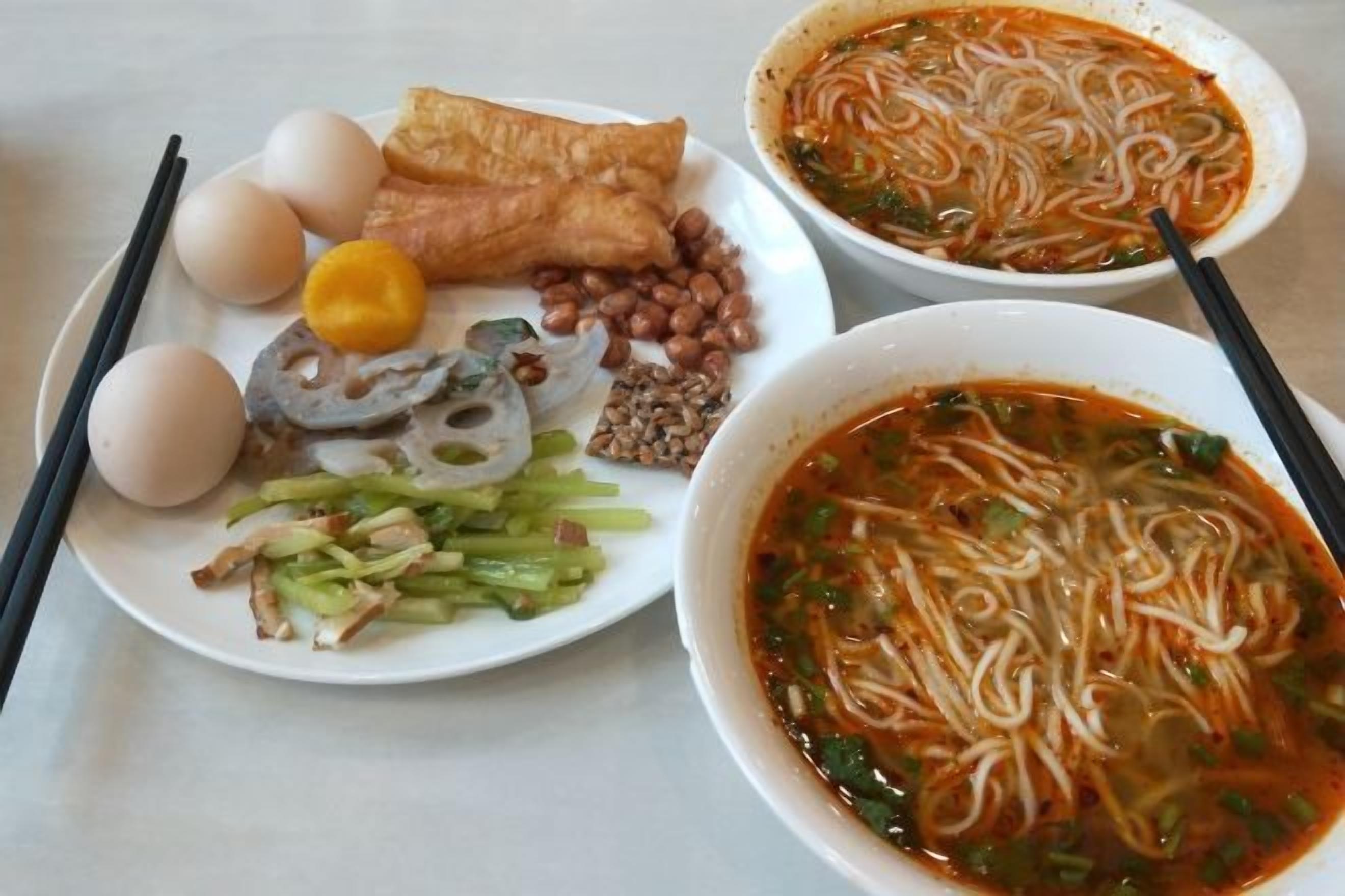 A table with two bowls of noodle soup and a plate with eggs, fish, and vegetables.