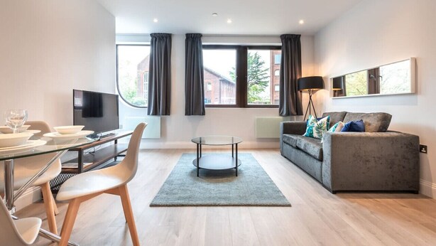 A modern living room with a glass dining table, a grey sofa, a round coffee table, and a flat-screen TV.