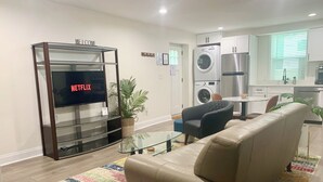 A modern living room with a leather sofa, a glass coffee table, and a colorful rug. A flat-screen TV is mounted on a glass cabinet labeled "WELCOME" and "NETFLIX". A kitchen with a washing machine and a refrigerator is visible in the background. - FIFA World Cup - 4Mi to Stadium - Private Basement Apt - Free Parking - Kitchen (Atlanta)