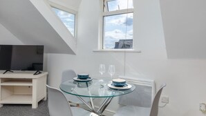 Una habitación pequeña y moderna con una mesa de comedor de vidrio, sillas blancas, un televisor sobre un mueble blanco y una ventana con vistas a edificios.