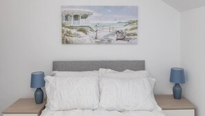 Un dormitorio con cama, dos mesitas de noche con lámparas y un cuadro colgado en la pared que muestra una playa.