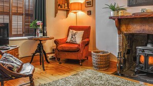 A cozy living room with a red armchair, a small table with a lamp, a fireplace, and a bookshelf with decorative items.