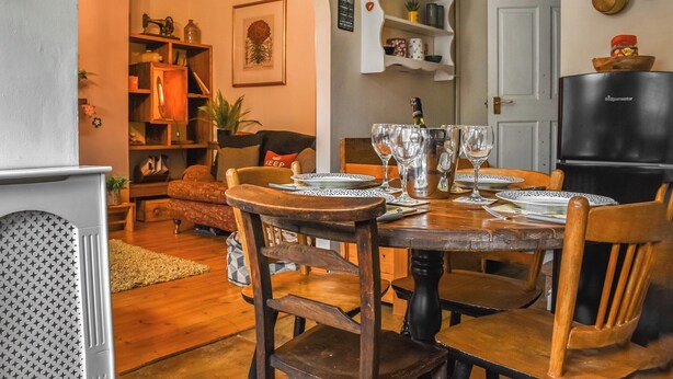 A cozy dining area with a wooden table set for four, surrounded by wooden chairs. A chandelier hangs above, and a bookshelf filled with various items is visible in the background.
